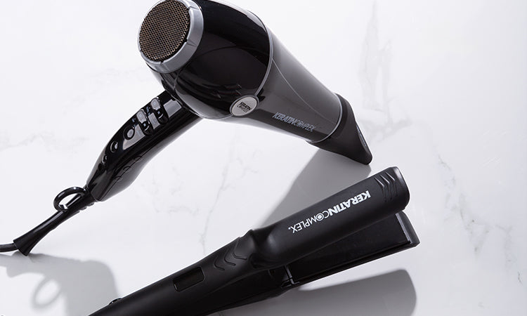 Keratin Complex black hairdryer and flat iron placed on a white marble surface.