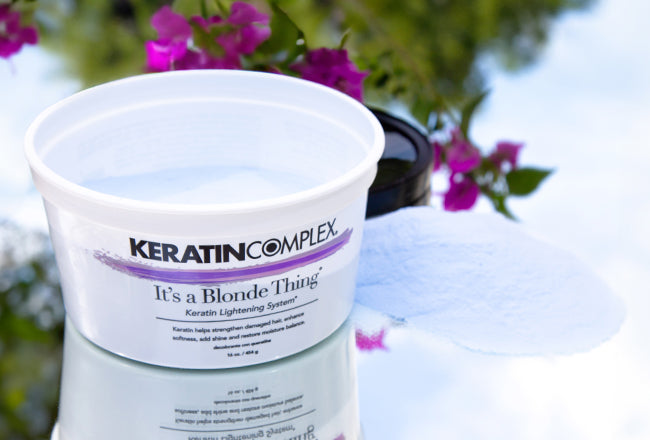 Keratin Complex 'It’s a Blonde Thing' Keratin Lightening System container, with lightening powder spilled beside it, set against a background of flowers.