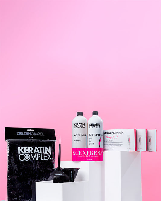 Keratin Complex hair treatment products displayed on white podiums against a pink background, including KC Primer, KCEXPRESS bottles, Vitalshot treatments, a Keratin Complex cape, and hair application tools (bowl and brush)