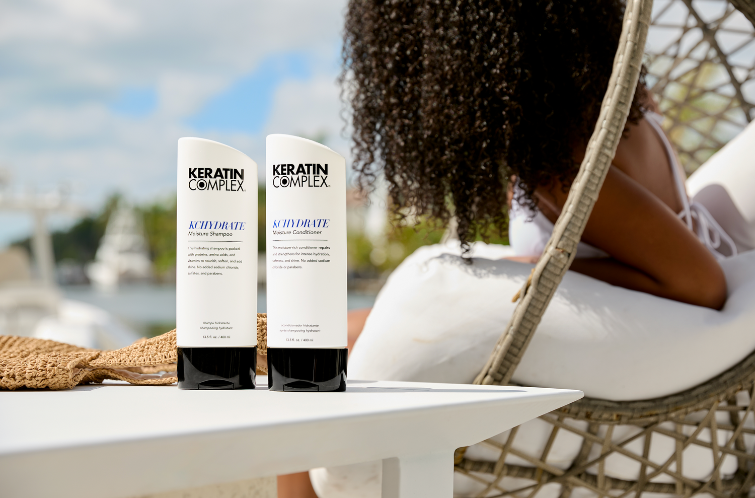 Two bottles of Keratin Complex KCTHYDRATE Moisture Shampoo and Conditioner are displayed on a white outdoor table next to a woven hat. In the background, a woman with curly hair is lounging on a cushioned wicker chair near a waterfront, with boats and greenery visible under a sunny sky.