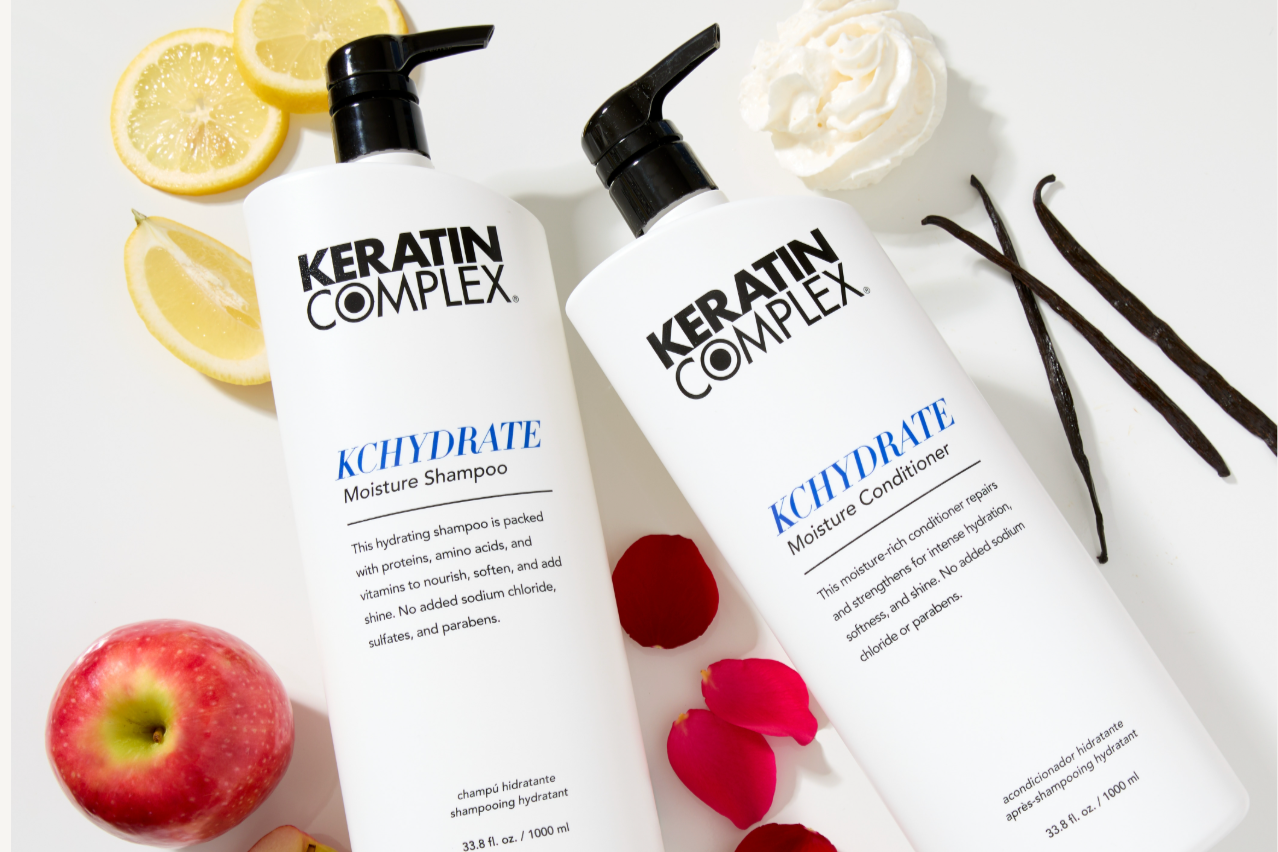 Two large pump bottles of Keratin Complex KCHYDRATE Moisture Shampoo and Conditioner are displayed on a white surface surrounded by fresh ingredients, including sliced lemon, a whole and sliced red apple, rose petals, vanilla beans, whipped cream.