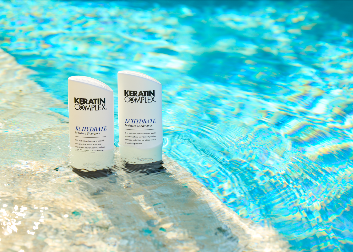 Two bottles of Keratin Complex KCHYDRATE Moisture Shampoo and Conditioner stand at the edge of a shimmering swimming pool, partially submerged in clear blue water, creating a refreshing and hydrating visual theme.