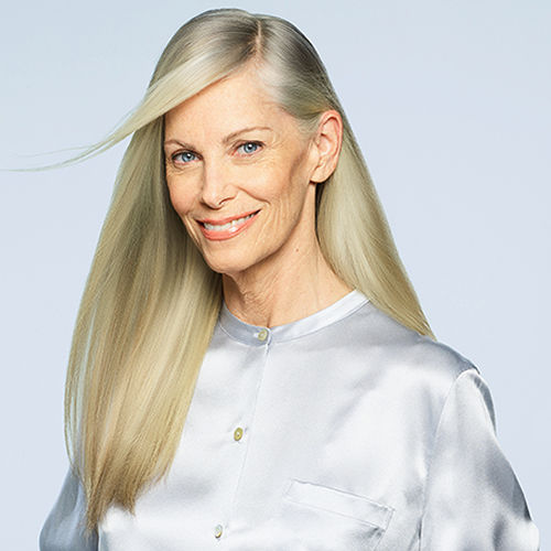A closeup of a woman who has long light blonde hair, she looks at the camera, she has on a silver shirt