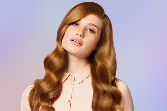 A closeup of a woman with long wavy red hair, she has on a light pink top, she is looking at the camera