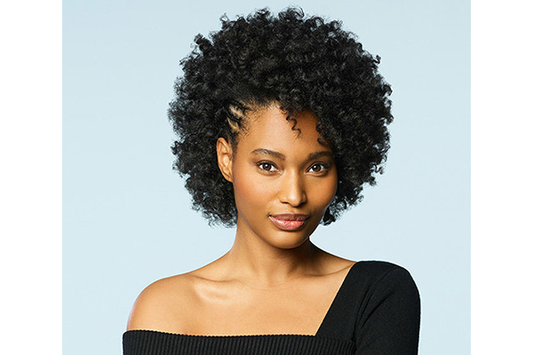 A closeup of a woman, she has short black curly hair, she looks at the camera and has on a black off the shoulder shirt