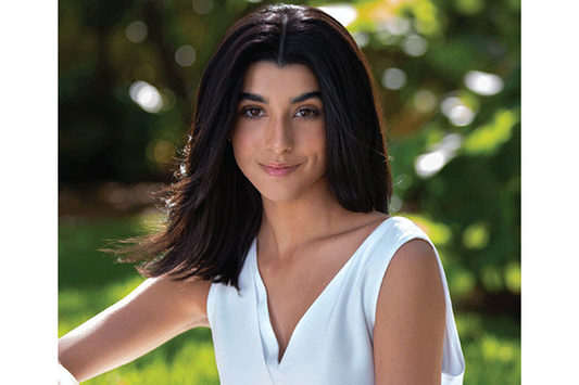 A waist up closeup of a woman who is outdoors, she looks at the camera, she has on a white shirt