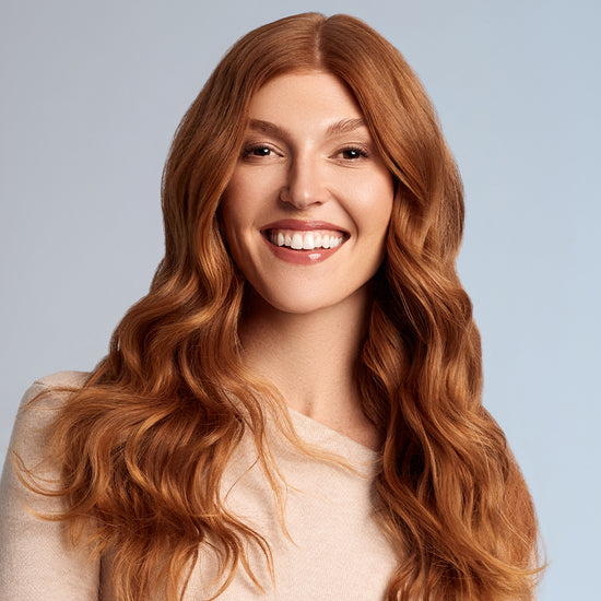 A person with long, soft waves of red hair, smiling and looking radiant in a beige sweater.