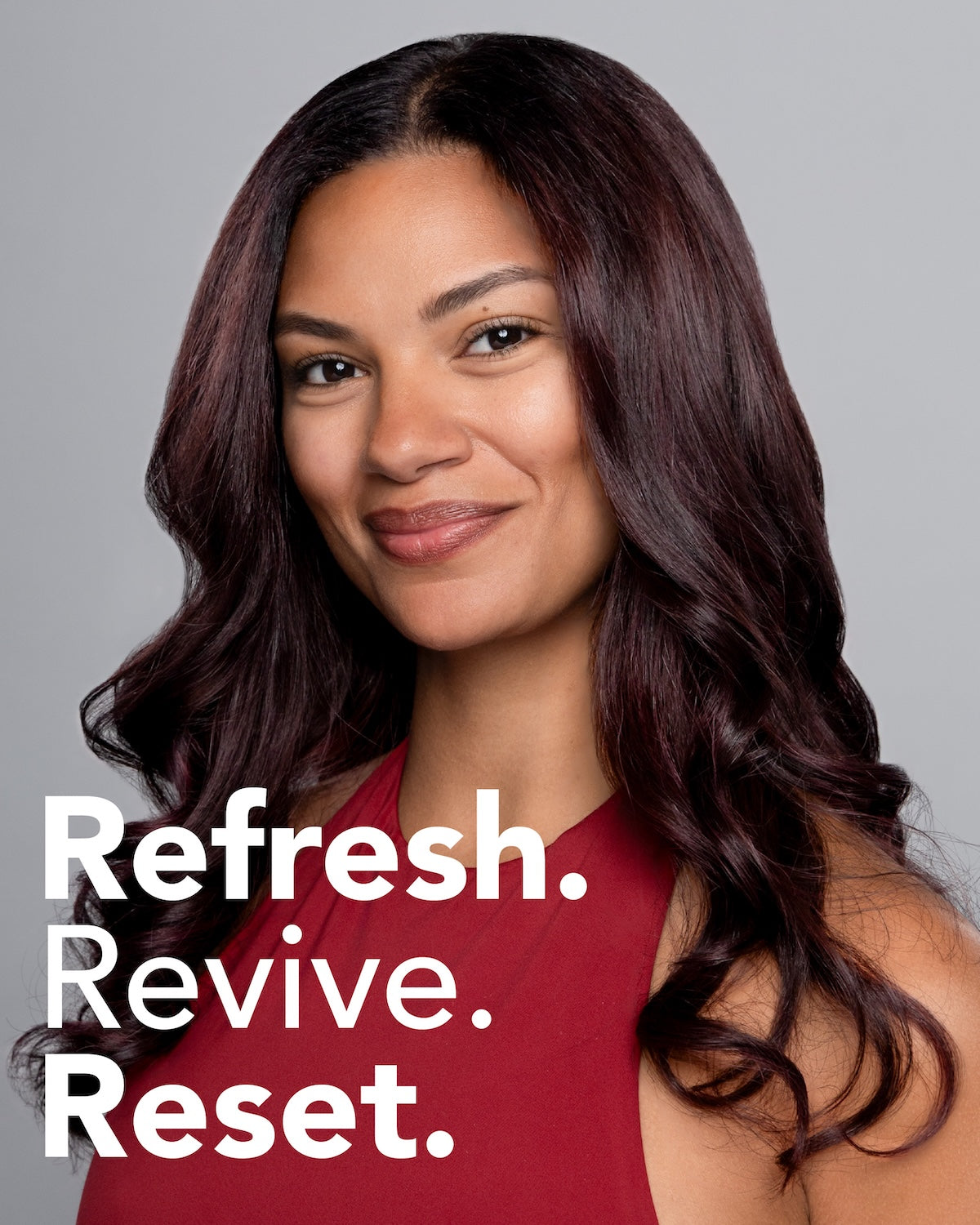 A woman with long, wavy dark auburn hair wearing a sleeveless burgundy top, set against a solid gray background ||Refresh. Revive. Reset.
