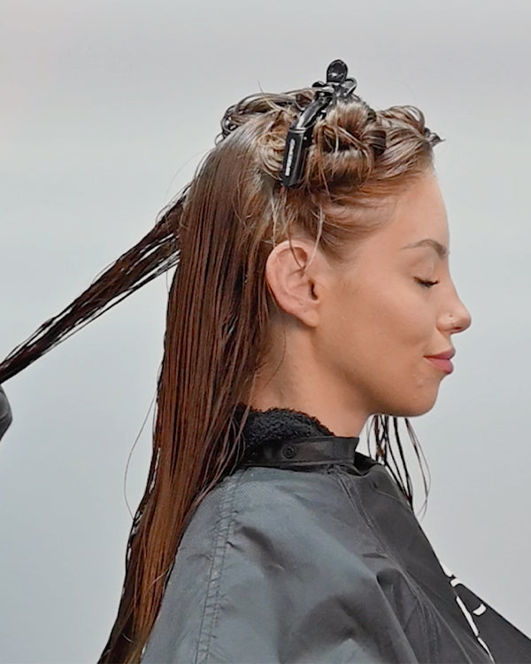 A woman with her hair sectioned and clipped, receiving a hair treatment while wearing a Keratin Complex cape in a salon.