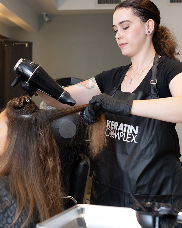 A stylist blow-drying a client's hair in a salon, wearing a Keratin Complex apron and black gloves, while using a round brush to style the hair.