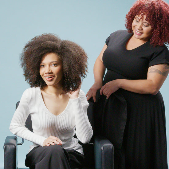 Smiling client with curly hair sitting in a salon chair, while the stylist stands beside her, both looking pleased with the result.