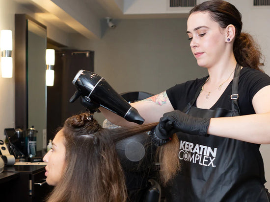 A hairstylist wearing gloves and a Keratin Complex apron blow-drying a client's hair in a modern salon setting.
