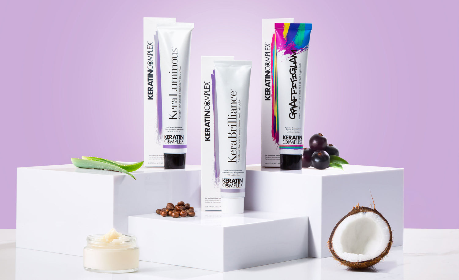 Three Keratin Complex hair color tubes—KeraLuminous, KeraBrilliance, and Graffitiglam—are displayed with aloe, coffee beans, acai berries, and a coconut, against a light purple background.