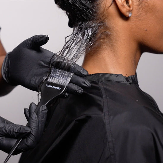 A close-up of a stylist applying a smoothing treatment to a client’s wet hair with a brush.