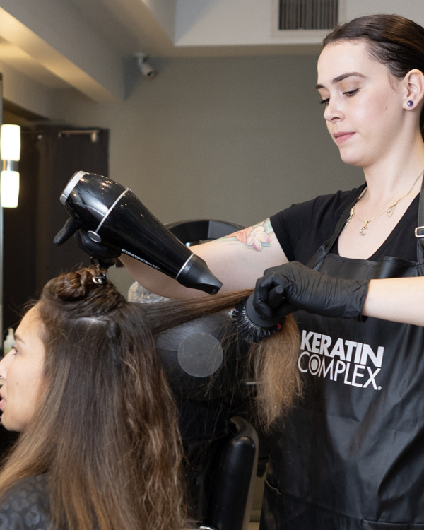 A stylist applying a hair treatment to a client's hair in a salon, wearing black gloves and a black outfit.