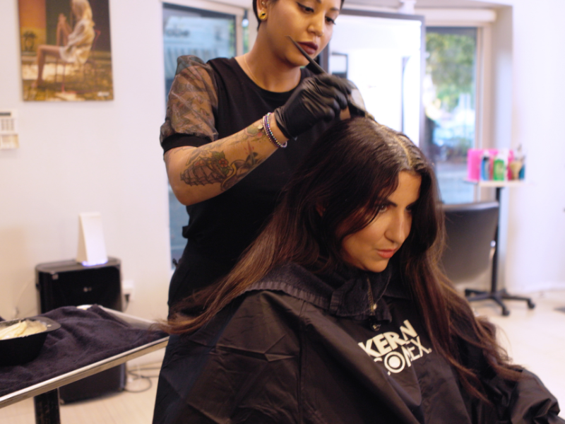 A hairstylist wearing black gloves and a black shirt with lace sleeves applies hair color to a seated client's roots.