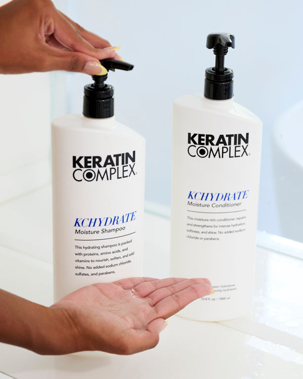 A close-up of a person's hands using the Keratin Complex KCHYDRATE Moisture Shampoo. One hand is pressing the pump of the shampoo bottle while the other is open, catching the product. Next to the shampoo is a bottle of Keratin Complex KCHYDRATE Moisture Conditioner. 