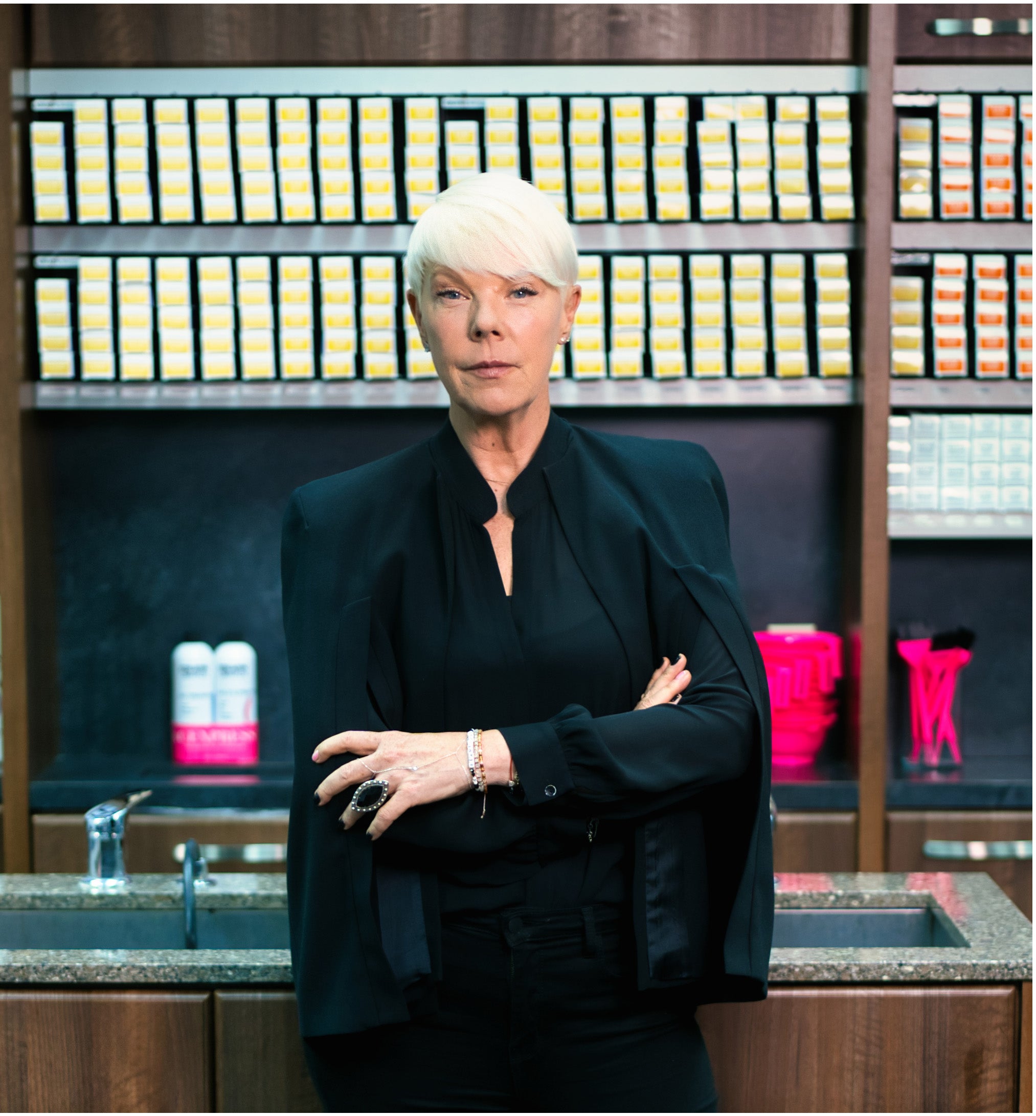 A woman with short white hair stands in front of a dark shelving unit filled with many small rectangular items, wearing a black satin blazer over a black top. The setting appears to be an indoor retail or studio environment with wood shelving and pink decorative objects in the background.