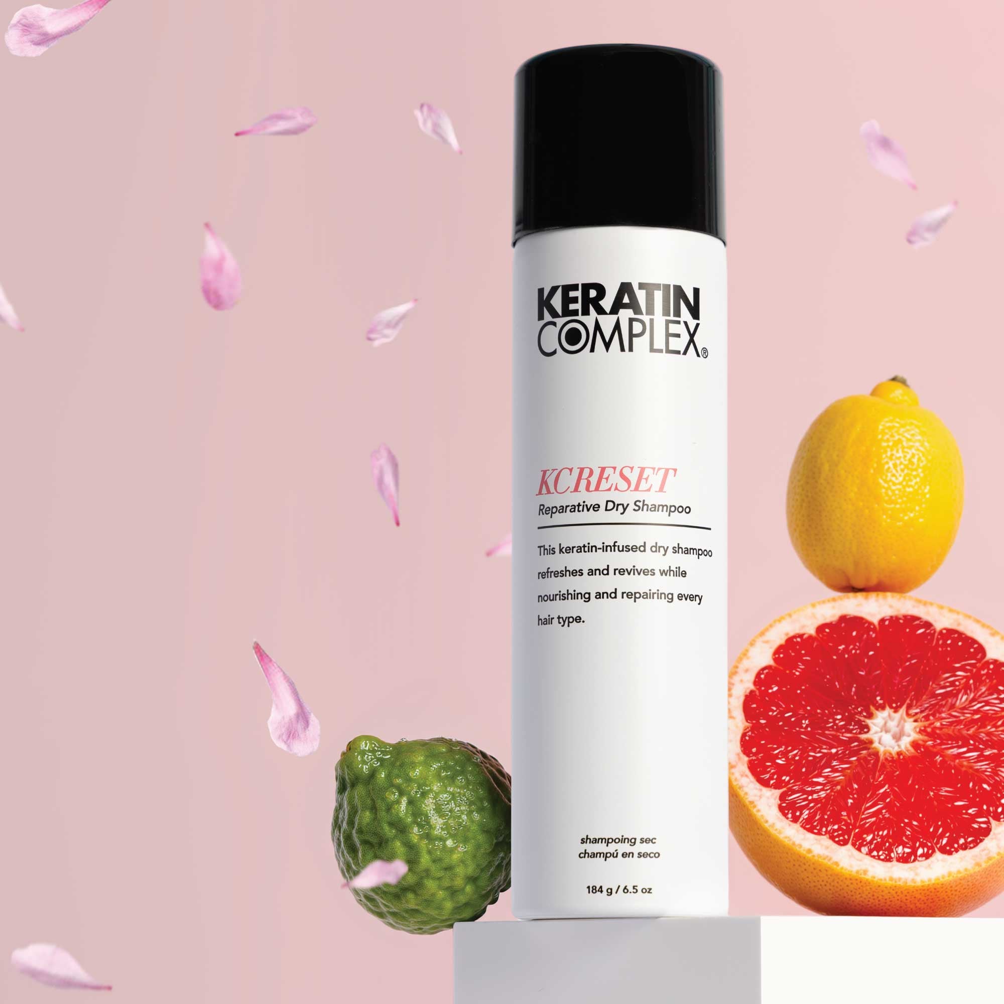 Keratin Complex KCREST, styled on a pedestal with a pink background and citrus fruits including a lemon, grapefruit, and a lime, with petals floating around.