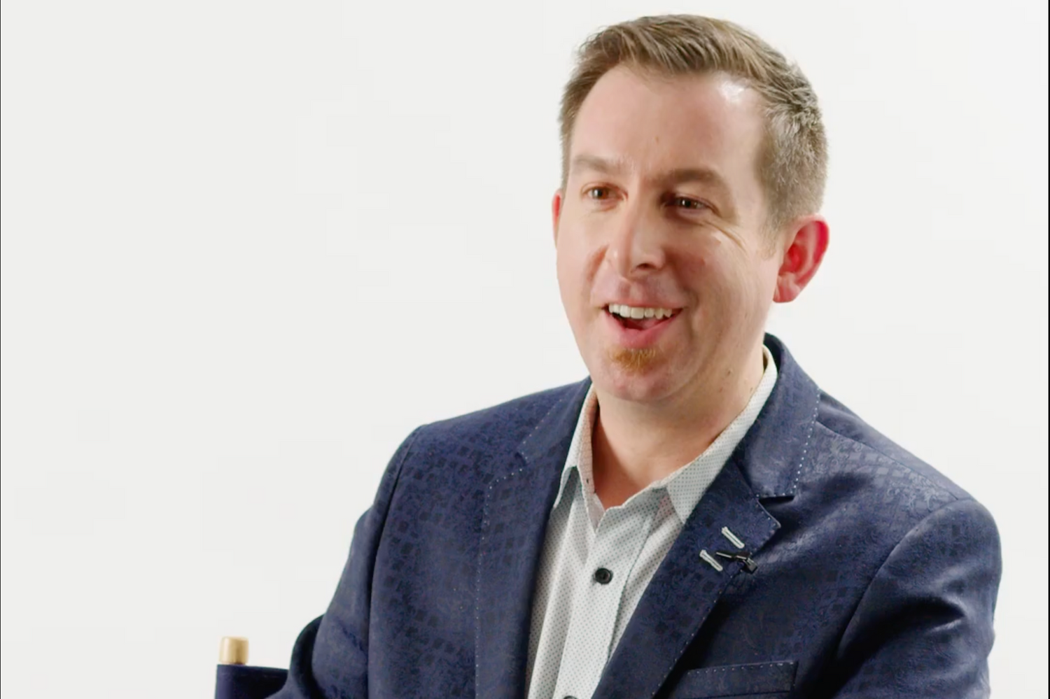 A man with short brown hair, wearing a navy blazer and gray shirt against a white background.