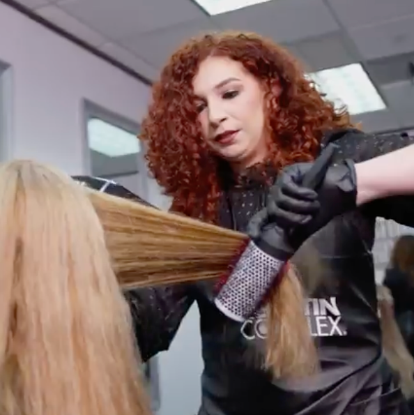A hairstylist with curly red hair uses a round brush and blow dryer to straighten a client's long blonde hair. 