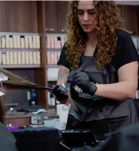 A hairstylist with long curly hair applies hair color to a client's hair using a brush.