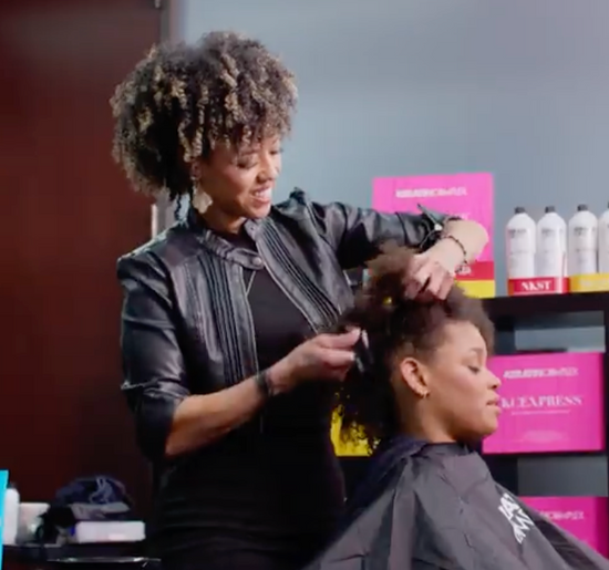A hairstylist in a black leather jacket styles a seated client's natural curly hair in a salon.