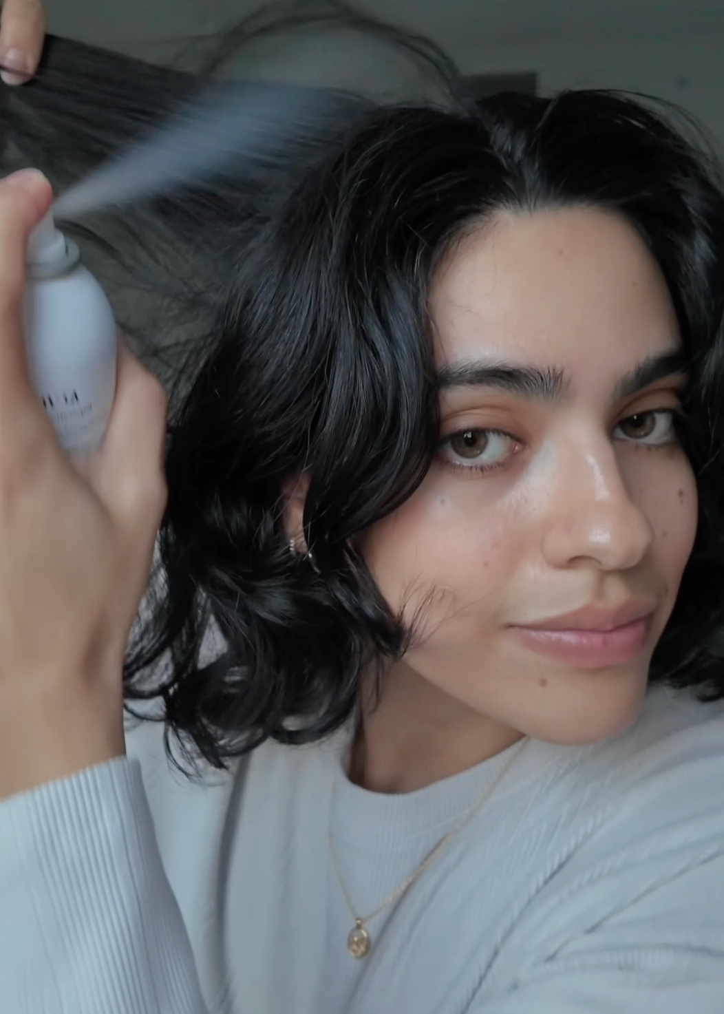 A woman with dark wavy hair sprays her hair with KCRESET.
