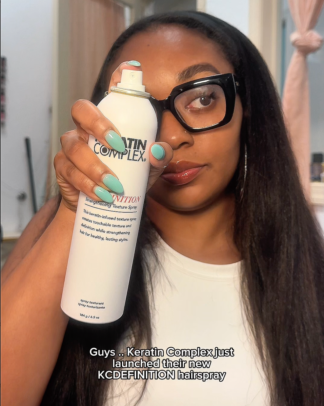 A woman with long black hair holds up a white can of Keren Complex KCDEFENITION Hairspray in front of her face.