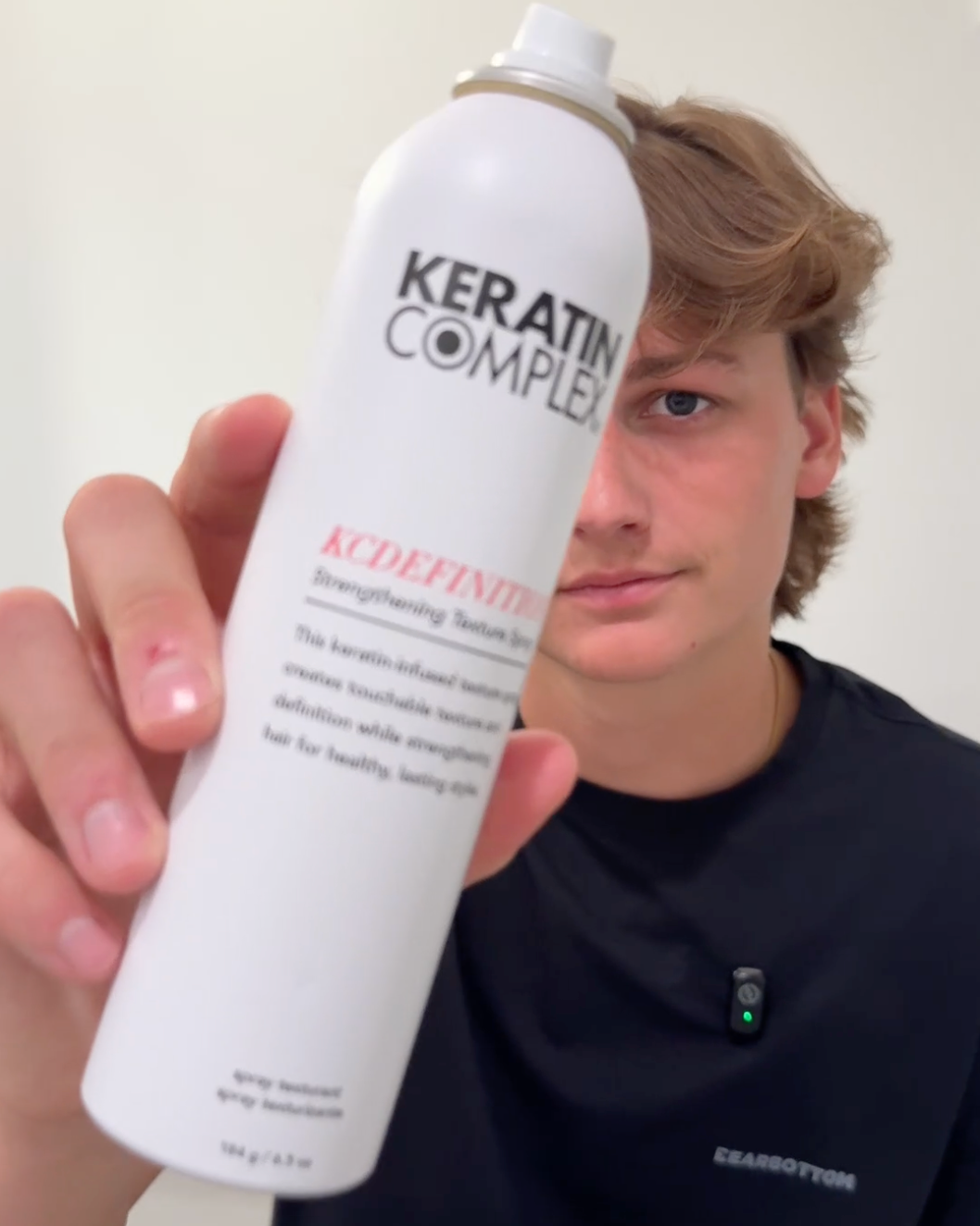 A person holding up a white can of Kerafin Complex KCDEFINTION Strengthening Hair Spray against a plain light background.